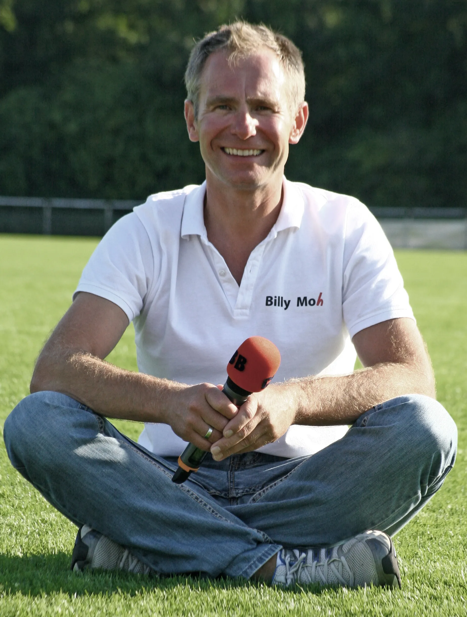 Sportmoderator Andreas Billy Moh sitzt lächelnd mit Mikrofon auf dem Rasen